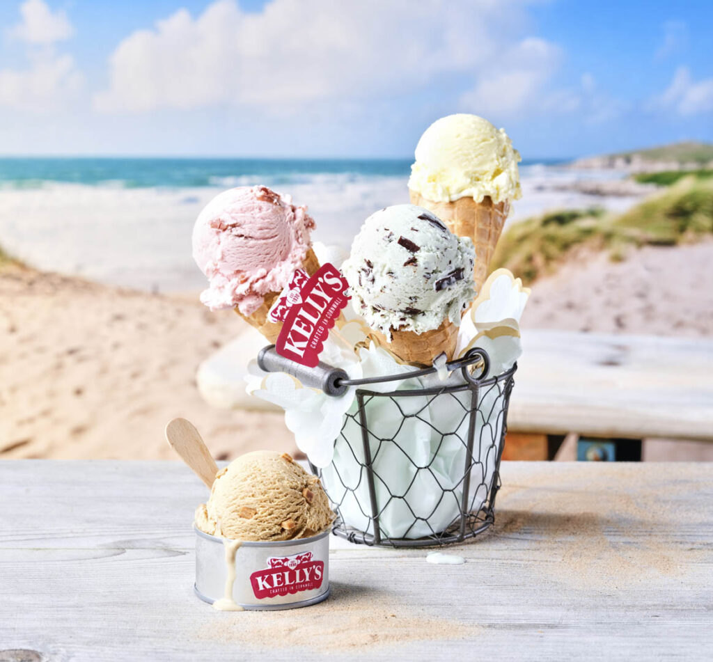 Three colorful ice cream cones in a bucket on a table, with a beach view, showcasing local Kelly's Ice Cream in Cornwall.