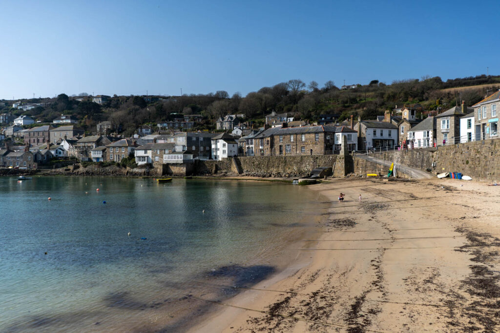 A sunny beach at Mousehole Harbour with a few people enjoying the sand and sea, the birthplace of the Stargazy Pie, a great Cornish recipe for Mother's Day.
