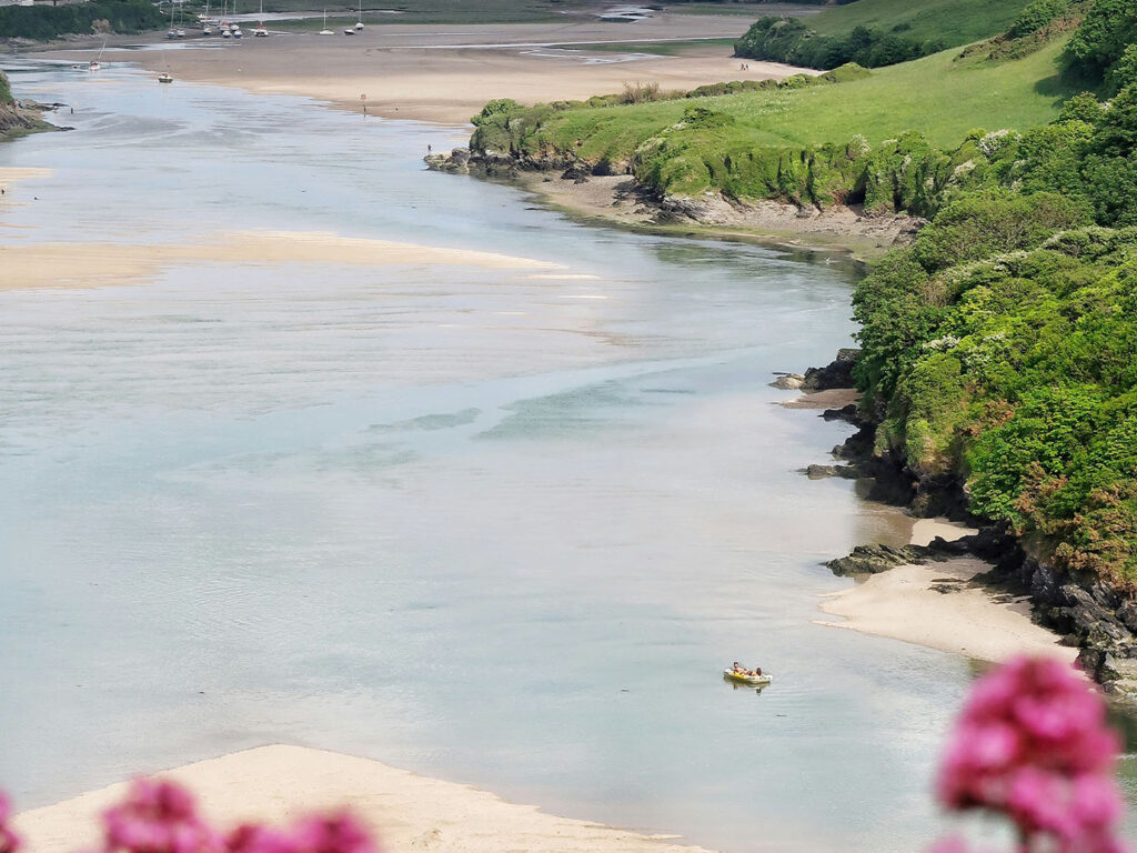 A serene river scene featuring a boat gliding through the Gannel Estuary in Newquay, surrounded by lush greenery, a great thing to do off the beaten track in Newquay.