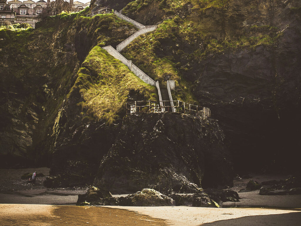 A scenic beach view featuring a staircase leading up to a cliff, showcasing Tolcarne Beach in Newquay, a great thing to do in Newquay off the beaten track.