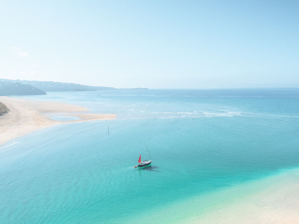 A sailboat with a red sail on the turquoise waters of Hayle Estuary, with a sandy beach and distant coastline, just one location in which Cornish Escapes can help with holiday cottage ownership.