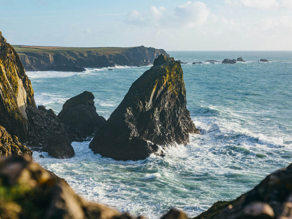 Stunning cliffs of Cornwall at Kynance Cove, showcasing rugged coastline and vibrant blue waters under a clear sky, a great place to visit from a Cornwall holiday cottage.