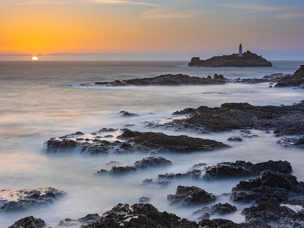 Godrevy Lighthouse stands tall at sunset, surrounded by rocky shores and gentle waves, casting a serene coastal scene, close to food and drink options in Hayle.