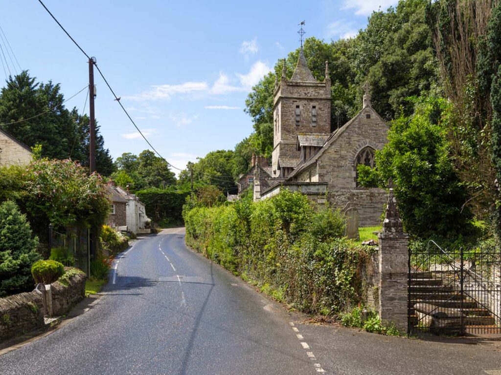 A scenic country road lined with trees, featuring a quaint church nestled beside it in Little Petherick village, a great piece of arts and heritage in Padstow.