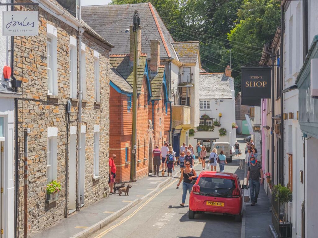 A bustling narrow street in Padstow, featuring pedestrians strolling and parked cars lining the sides, with a Rick Stein shop, central to Padstow's food and drink scene.