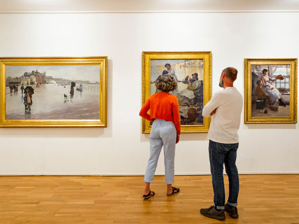 Two visitors admire paintings at Penlee House, surrounded by vibrant artwork and a warm atmosphere, a key part of Mount's Bay's arts and heritage.