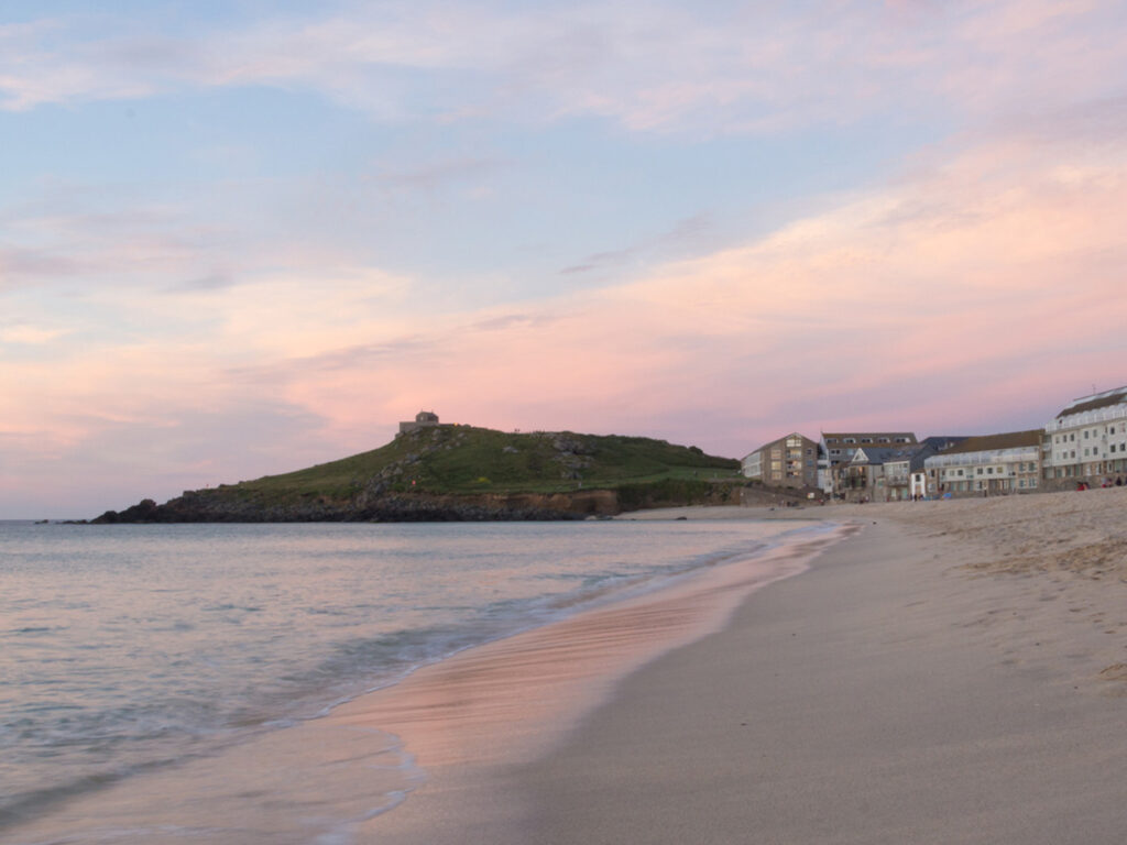 A picturesque view of Porthmeor Beach in St Ives, under a clear blue and pink sky, a part of St Ives' art and heritage scene.