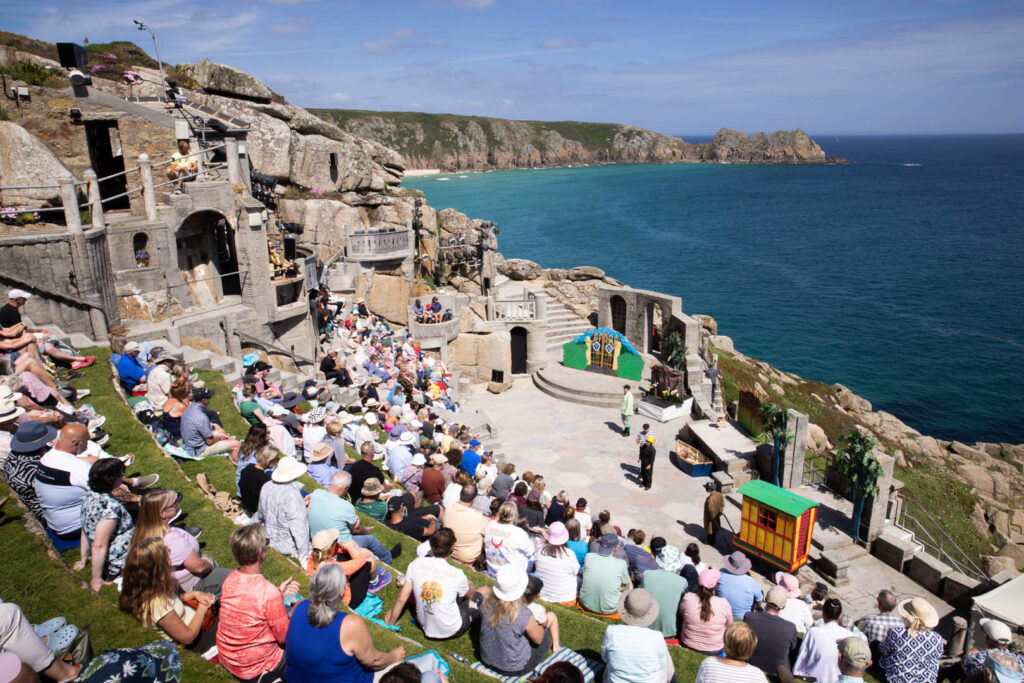 A lively crowd enjoying a performance at Minack Theatre, surrounded by stunning coastal views, a great thing to do in Cornwall with the kids this Summer.