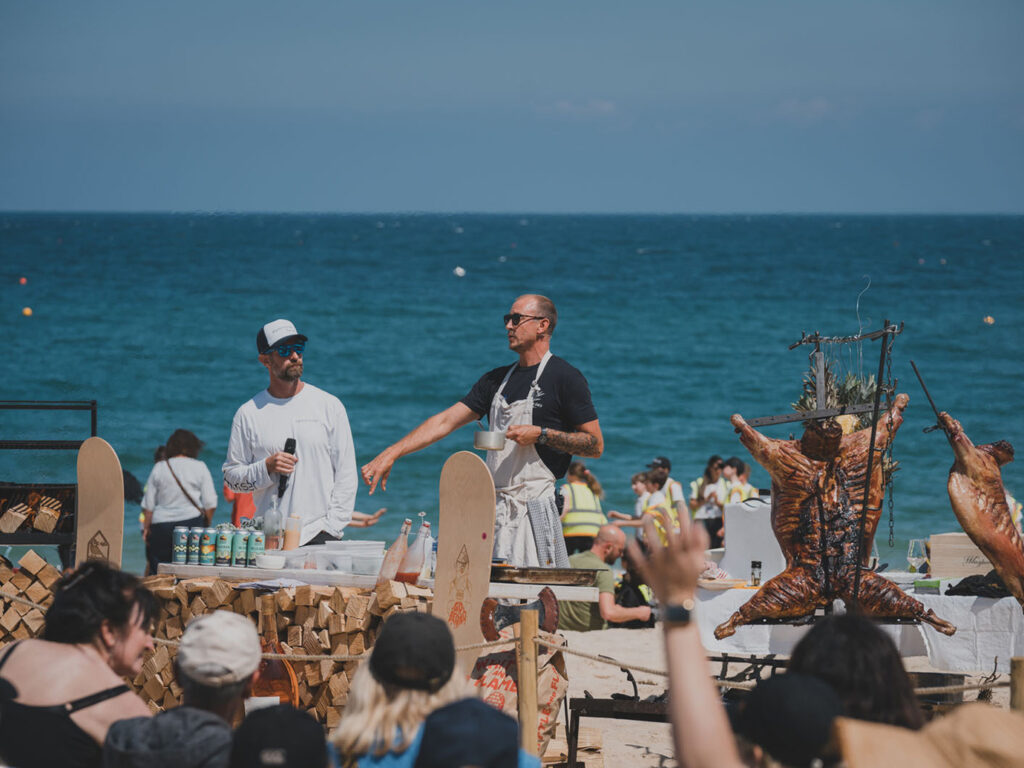 Two chefs demonstrate cooking on a beach during the St Ives Food Festival, with an audience watching eagerly, don't miss out on all of the food and drink.