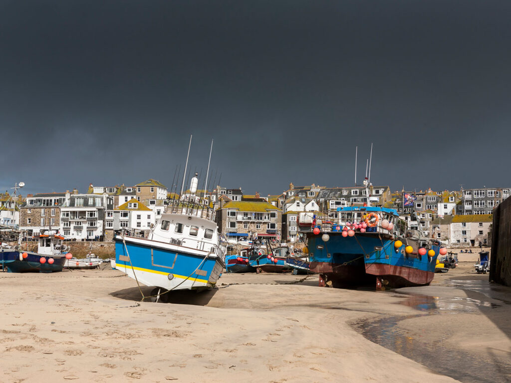 A moody sky looms over St Ives Harbour, where vibrant fishing boats are anchored, creating a picturesque scene, fishing was a key part to St Ives heritage.
