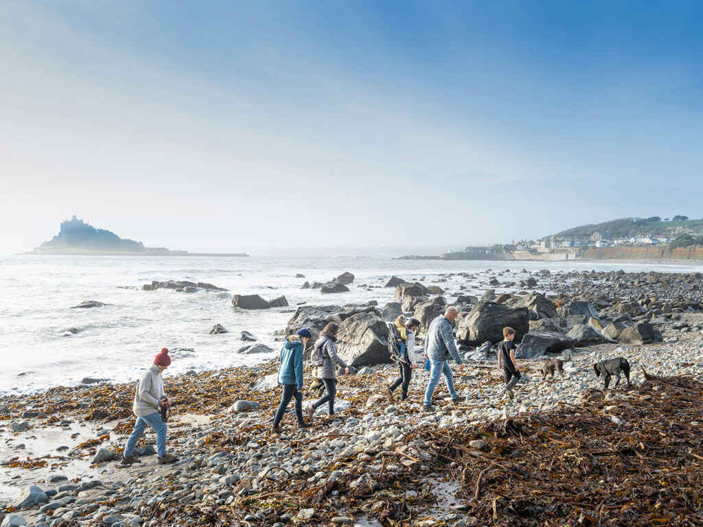 A group of people strolls along Marazion Beach, enjoying the sun and walking a happy dog by the shoreline, close to many food and drink options in Marazion.