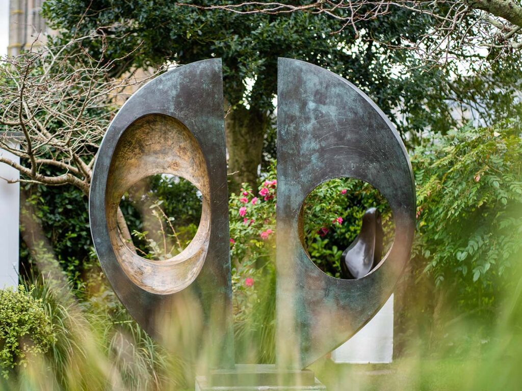 Large Barbara Hepworth sculpture, surrounded by greenery and a tree in the background, showcasing artistic heritage in St Ives.