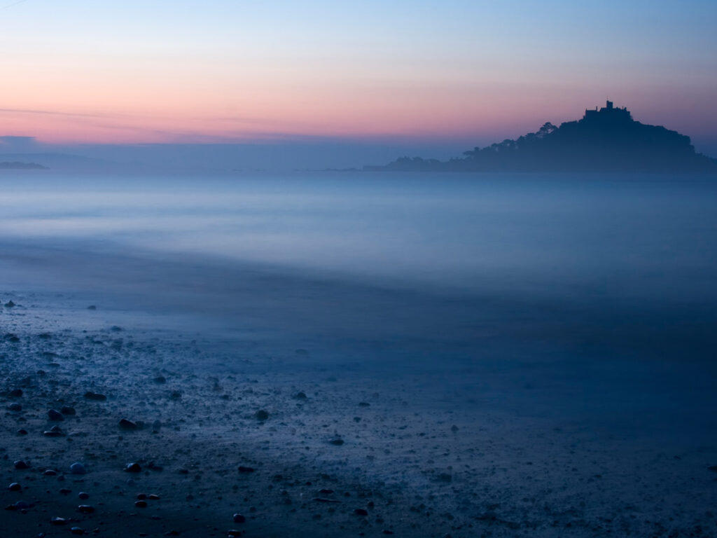 Long exposure of St Michael's Mount at sunset, with vibrant colors reflecting on the calm beach waters, a key part to Mount's Bay's arts and heritage scene.