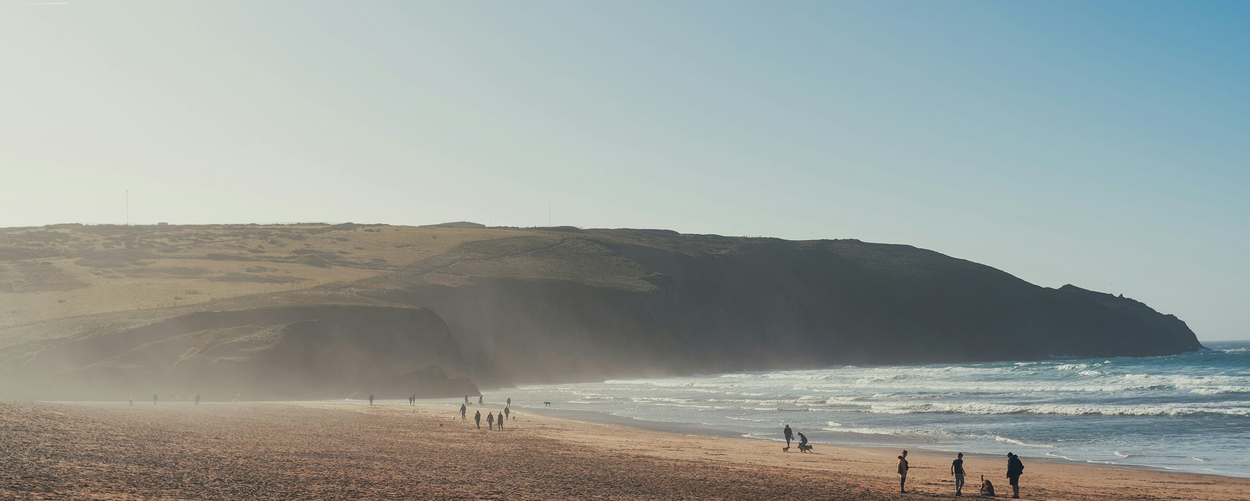 A sunny beach in Cornwall with people strolling along the shore, enjoying the warm weather and scenic views, a great place to visit from a cottage in Cornwall.