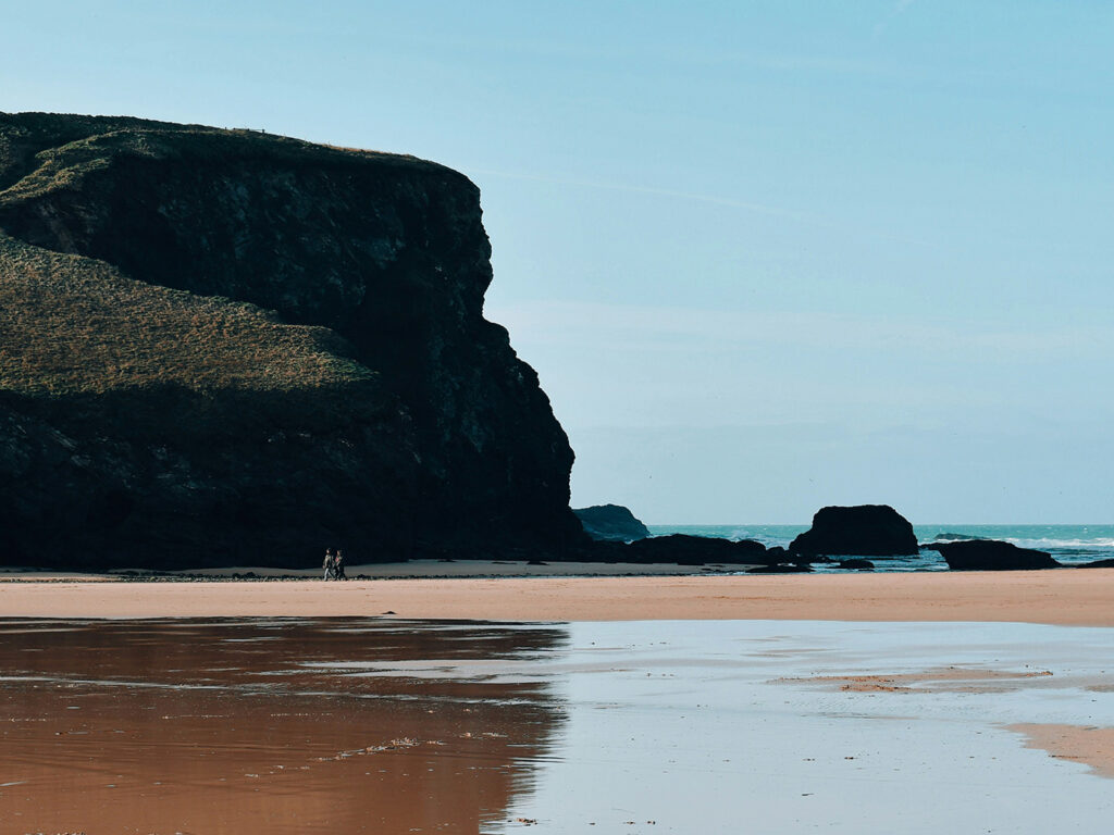 A person strolls along Mawgan Porth Beach, with waves gently crashing in the background, a place with lots of food and drink options.