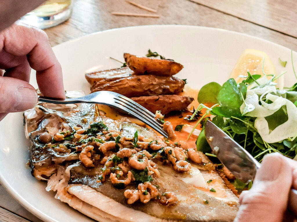 A person cuts a piece of fish with a fork, enjoying a seafood dish at Mawgan Porth Restaurant near Newquay, a great place for food and drink.