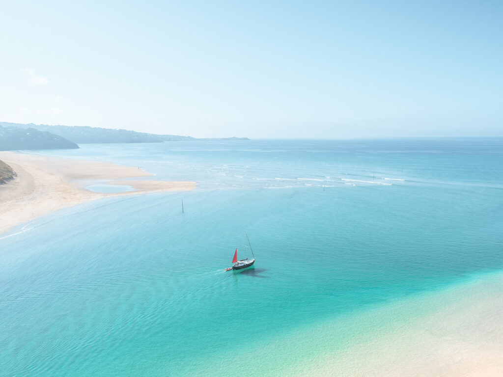 A boat gently floats in the Hayle Estuary, with a sandy beach visible in the background under a clear blue sky, moments from many Hayle food and drink options.