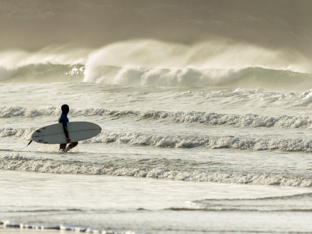 A man walks through the water at Fistral Beach, holding a surfboard, ready to catch some waves in Newquay, a great place for food and drink.