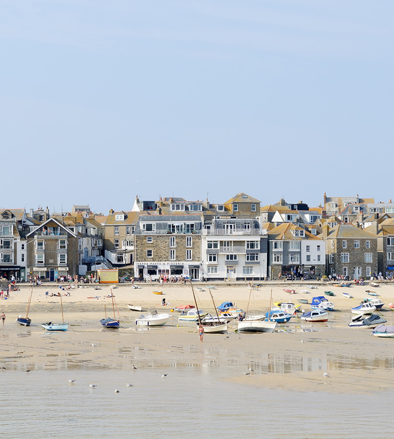 A sunny St Ives Harbour scene featuring a sandy beach dotted with colourful boats and gentle waves lapping the shore.