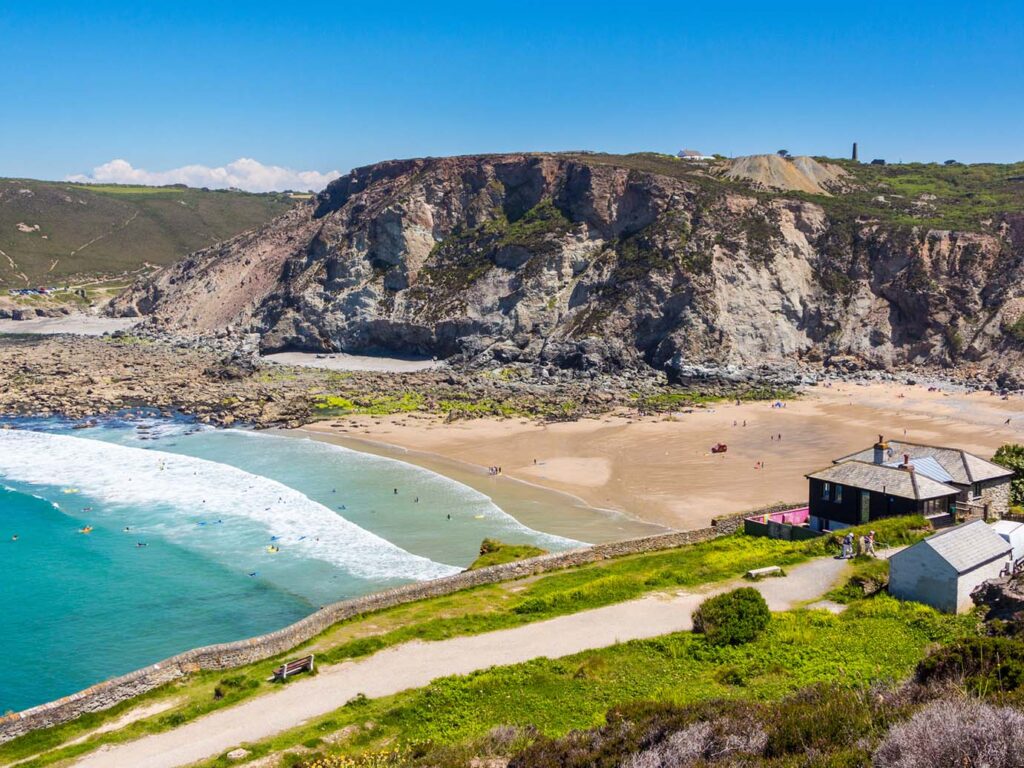 A picturesque view of Trevaunance Cove beach, featuring a cozy house perched on the sandy shore against a backdrop of cliffs, don't miss out on St Agnes food and drink nearby.